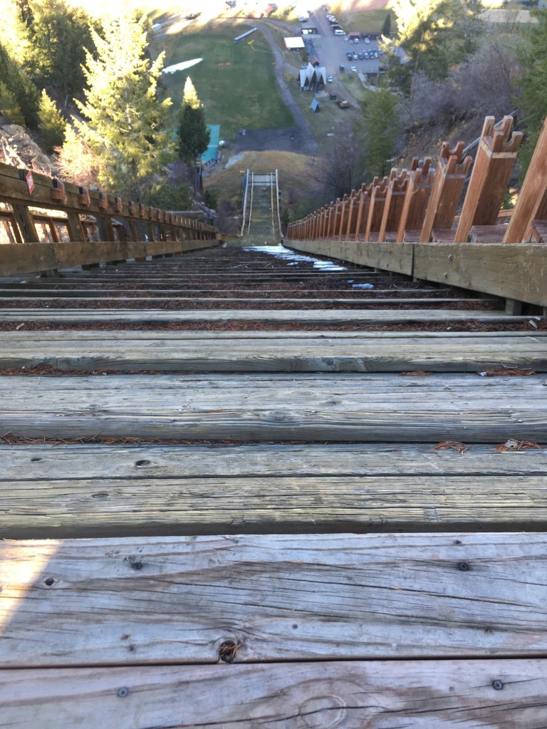 photo looking down from the top of a wooden ski jump platform to the lip of the jump and the landing hill below.