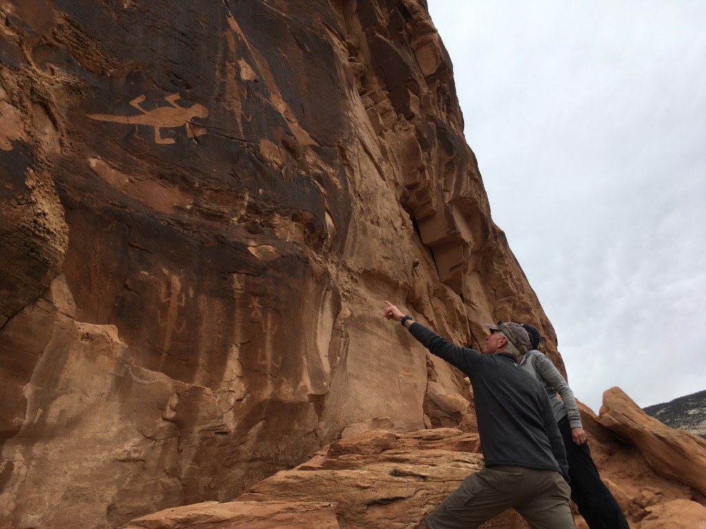 photo of two people pointing at a cliff with lizard petroglyphs