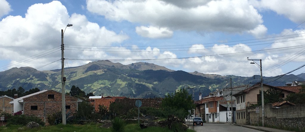 photo of green mountains, blue sky with puffy white clouds, and in the foreground, two-story brick houses with red tile roofs behind telephone poles and wires