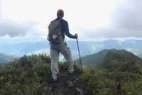 Photo of a male hiker standing atop a ridge overlooking a vast panorama of green mountain peaks just below the clouds.