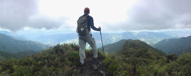 Photo of a male hiker standing atop a ridge overlooking a vast panorama of green mountain peaks just below the clouds.