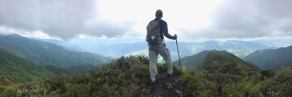 Photo of a male hiker standing atop a ridge overlooking a vast panorama of green mountain peaks just below the clouds.