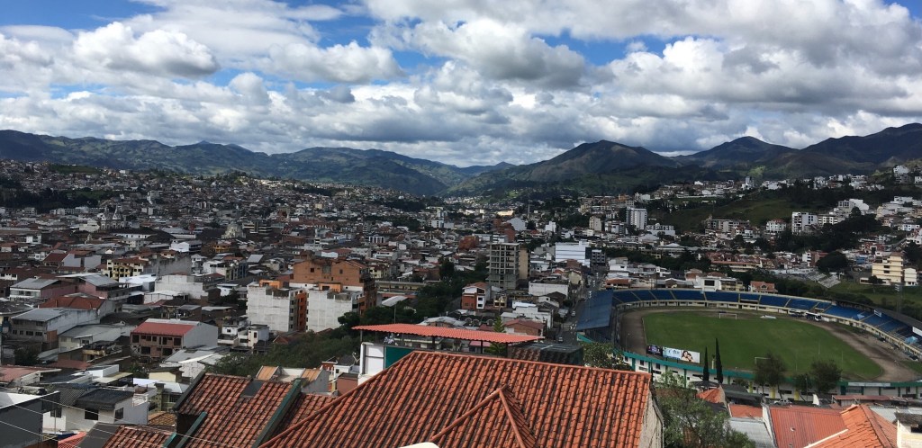 photo of the city of Loja, in a valley surrounded by mountains, with red tile roofs and a soccer stadium.