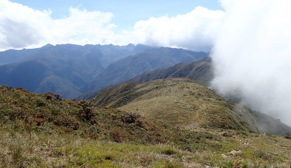 Photo of clouds sweeping up from the right to the top of a green mountain ridge, with green Andes mountaintops and blue sky to the left.