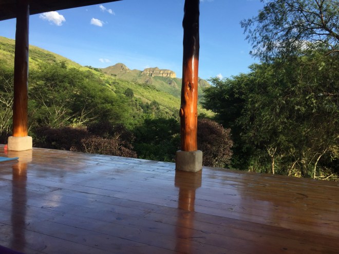 photo of a distant stone ridge, blue sky, green hills, taken from inside an open air, wood floor yoga studio with deep golden brown wooden pillars