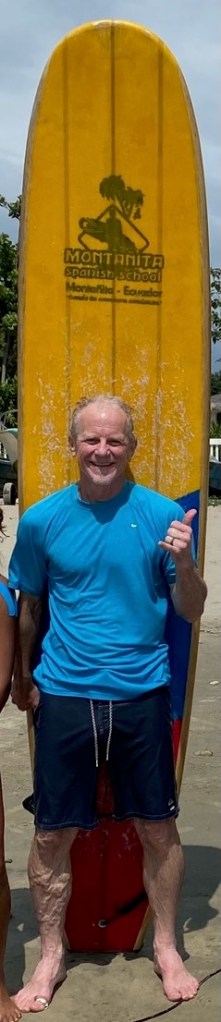 photo of Al (the author) in a blue swim shirt and bathing trunks standing in front of a tall yellow surfboard