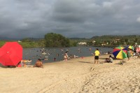 Photo of a beach in the foreground with people surrounding a still lagoon and colorful umbrellas, while dark clouds gather on the horizon.