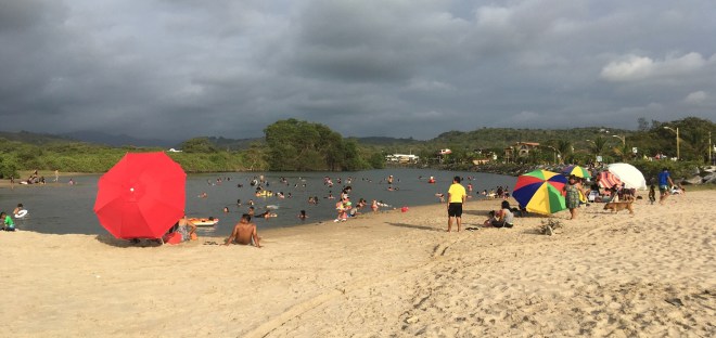 Photo of a beach in the foreground with people surrounding a still lagoon and colorful umbrellas, while dark clouds gather on the horizon.