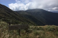 photo of a sign in Spanish, saying "Precaucion no salir del sendero" with a steep, green mountain ridge in the background.