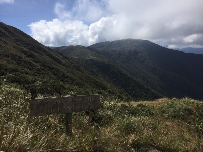 photo of a sign in Spanish, saying "Precaucion no salir del sendero" with a steep, green mountain ridge in the background.