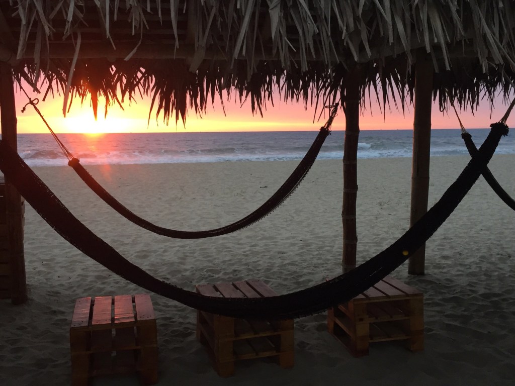 Photo of a beach sunset, seen through a palapa (beach hut) with two hammocks.