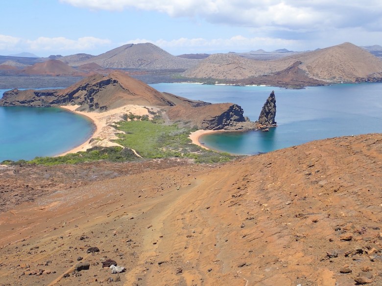 photo of a rock spike poking up out of green-blue water next to brown rocky hills and a yellow crescent beach, with red rocks in the foreground and brown hills in the background.
