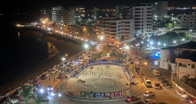 photo of a street at night beside a harbor, crowded with cars and people, winding around a roundabout with buildings and bright lights
