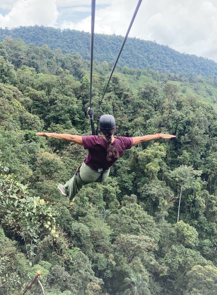 Photo of a woman wearing a helmet, maroon shirt, and green pants, hanging from a cable high above trees, with her arms outstretched as if flying.