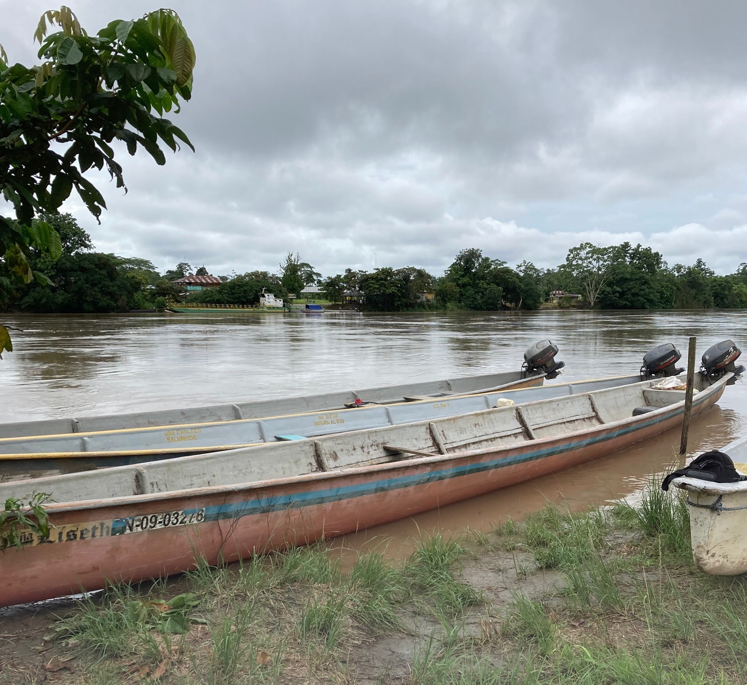 photo of three long canoes with outboard motors pulled up side by side on the edge of a wide brown river with leafy trees behind