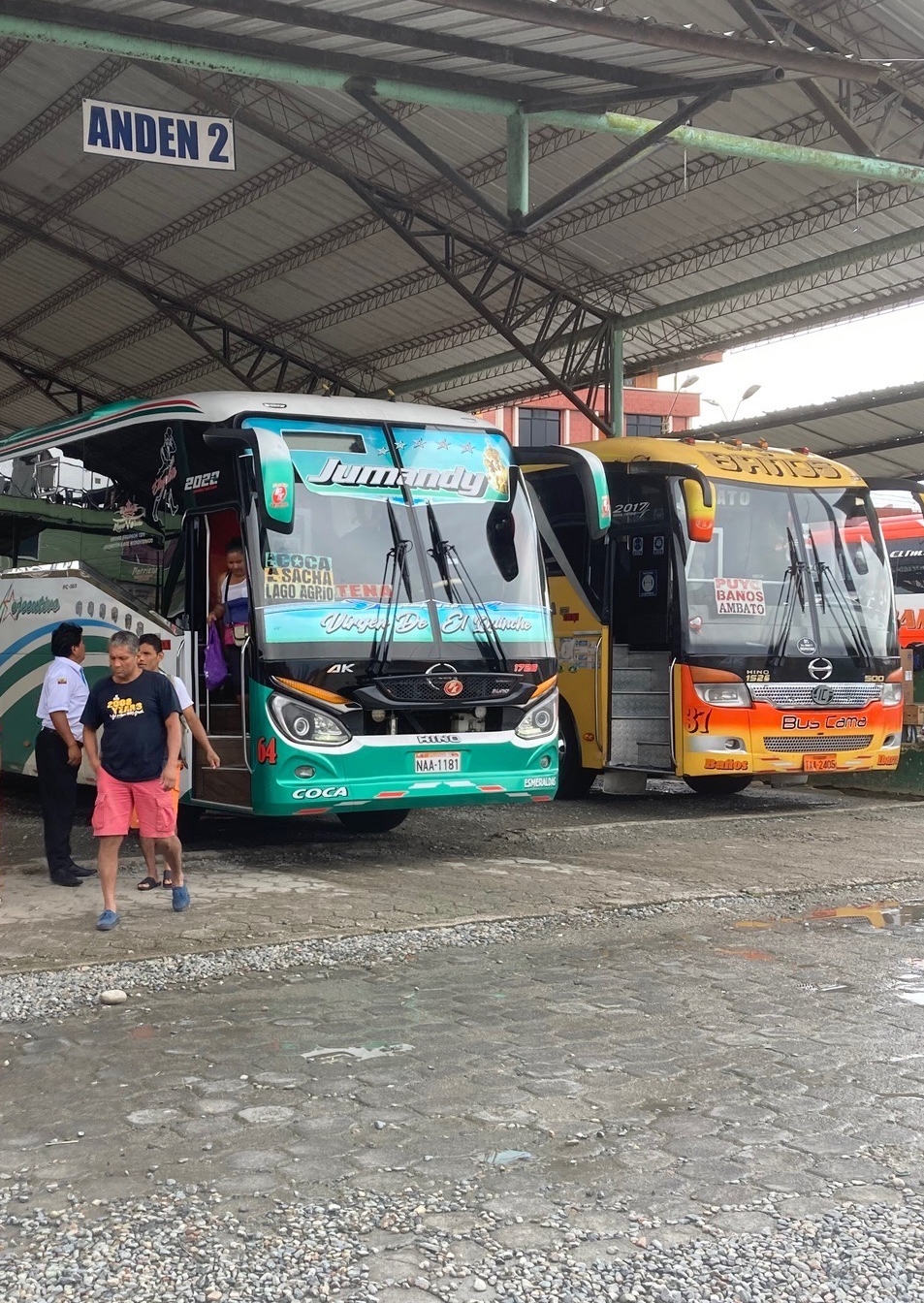 photo of a bright teal bus parked next to a bright orange bus under a roof with a sign above reading "ANDEN 2"