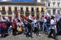 photo of a crowd of people waving white and red flags, some in traditional Andean clothes, marching in a disorderly parade across a cobblestone courtyard in front of colonial-style buildings.
