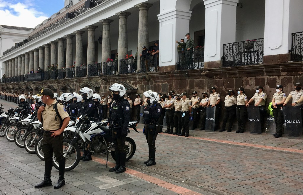 photo of a long line of police holding riot shields and motorcycles at the edge of a brick courtyard against a long building with white arches.
