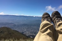 Photo of legs and feet in tan pants and hiking boots pointing up to the sky on the right side, with a green hill on the left and a distant cityscape far below.
