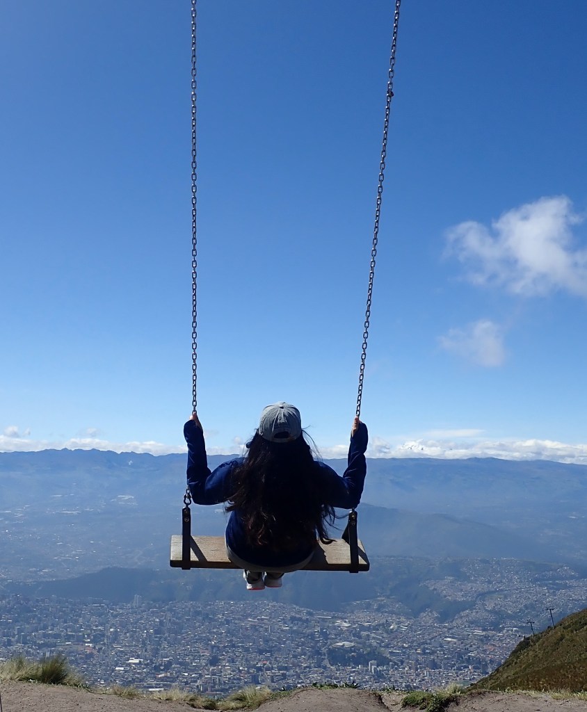 Photo of a woman on a large, flat-based swing, appearing to be out over the edge of a cliff overlooking a city far below and mountains in the distance.