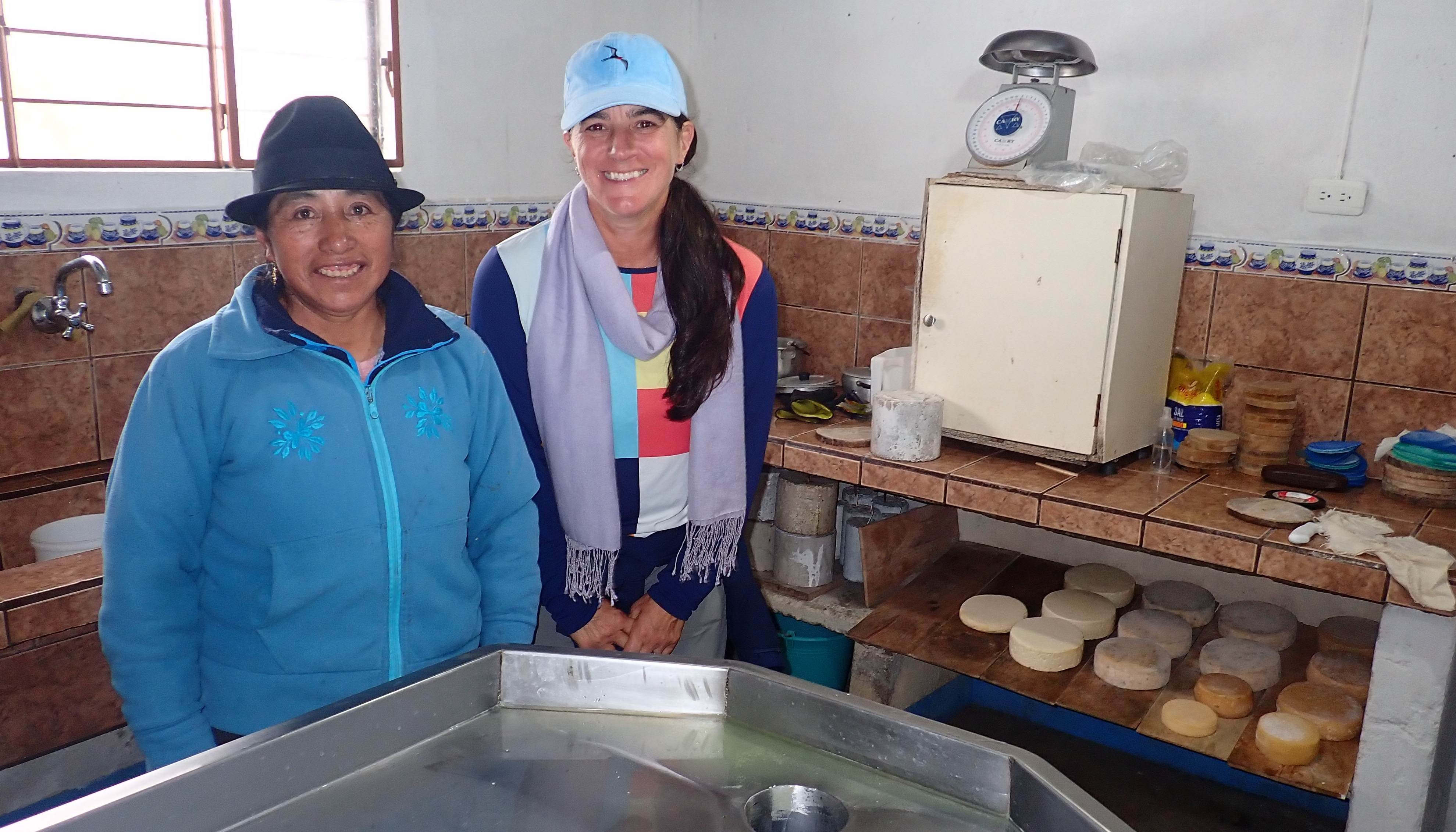 photo of two women, one in a teal sweatshirt and dark fedora, the other in a multi-colored hiking shirt, violet scarf, and blue cap, standing in a kitchen with wheels of light yellow cheese in the background.