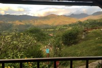 Photo of a sunlit mountain range in the distance, past a wide green valley, with a porch deck railing and roof in the foreground.
