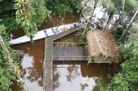 Photo from above, looking down on a long, narrow, white canoe next to a brown dock attached to a thatched-roof hut, sitting on reflecting, brown water and surrounded by leafy trees.