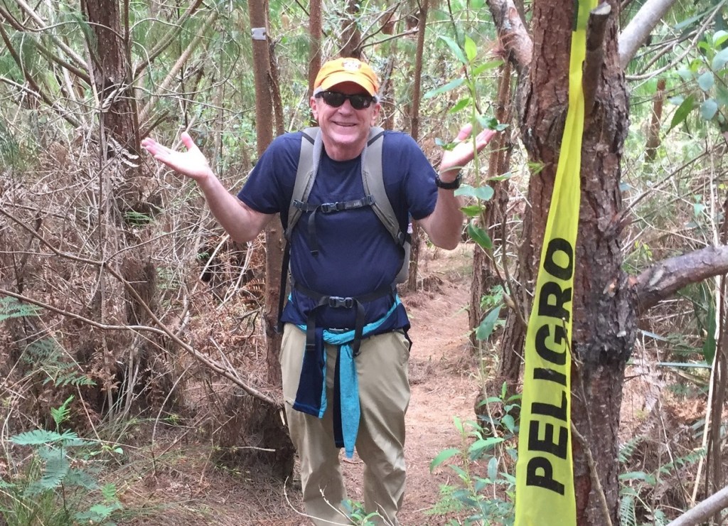 Photo of Al in a blue t-shirt, tan pants, orange baseball cap and dark sunglasses, standing on a hiking trail amidst trees, holding his hands upturned in a "who knows?" gesture, with a yellow tape reading "Peligro" ("danger" in Spanish) dangling from a tree in the foreground.