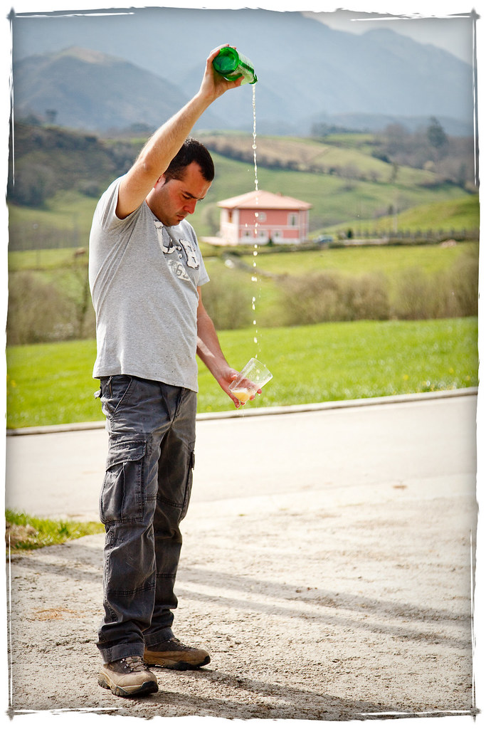 photo of a main pouring cider from a green bottle above his head into a glass by his waist, against a backdrop of green mountains.