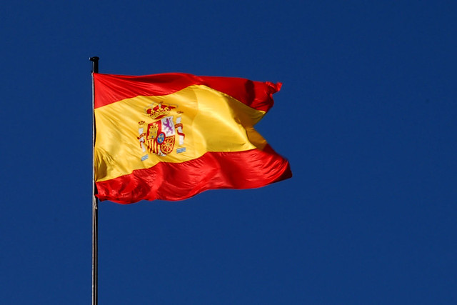 photo of red-and-yellow flag of Spain on a flagpole, waving in a wind, against a dark blue sky