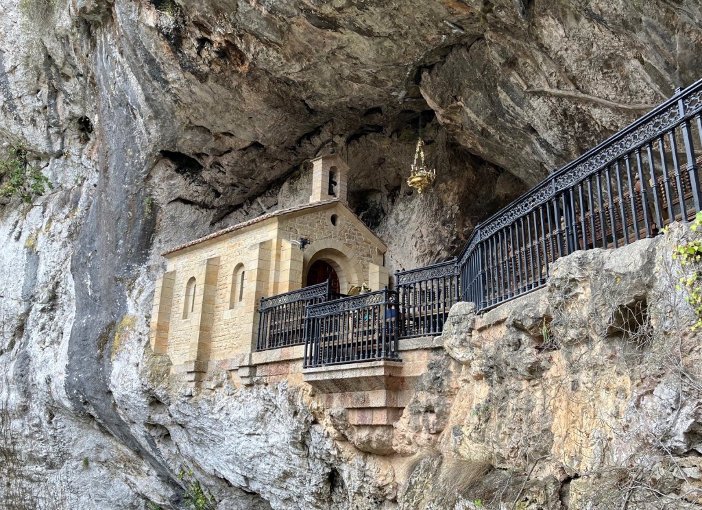 photo of a sandy-colored chapel with two windows, built into an open-sided cave on a cliff, with a black iron fence along the cliff.