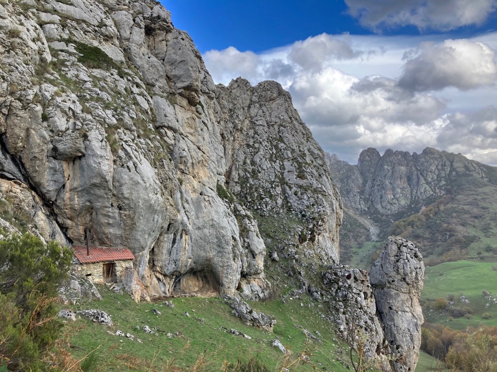 photo of a steep gray cliff with a small stone shelter with a red roof built into its base, with more cliffs and green fields in the distance.