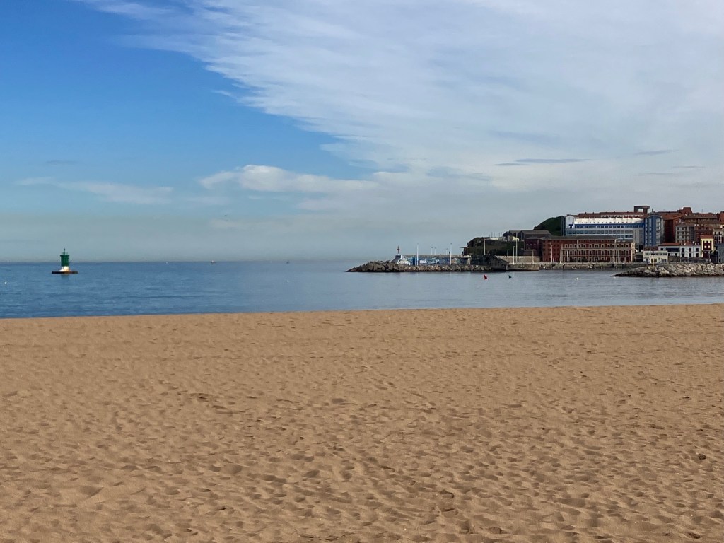 photo of a beach with tan sand and no people on it, with water and a rocky point with brick buildings and sailboat masts in the background.