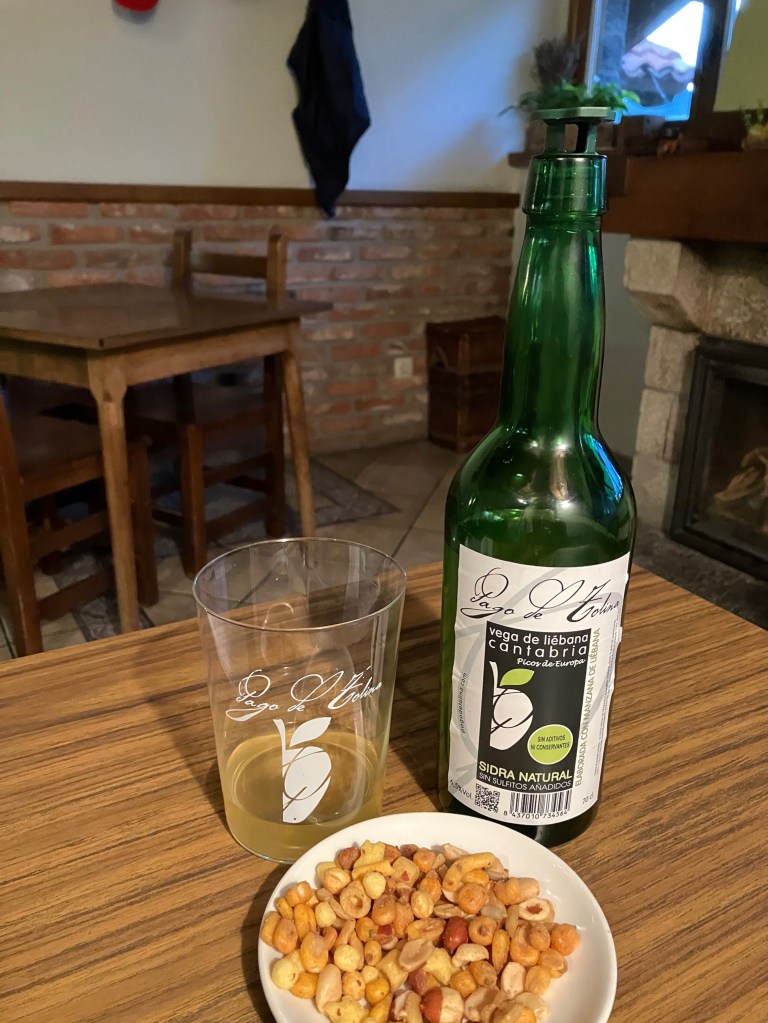 photo of a green bottle of Cantabrian cider next to a glass with cider in it and a plate of nuts in front, on a table in a brick-walled room