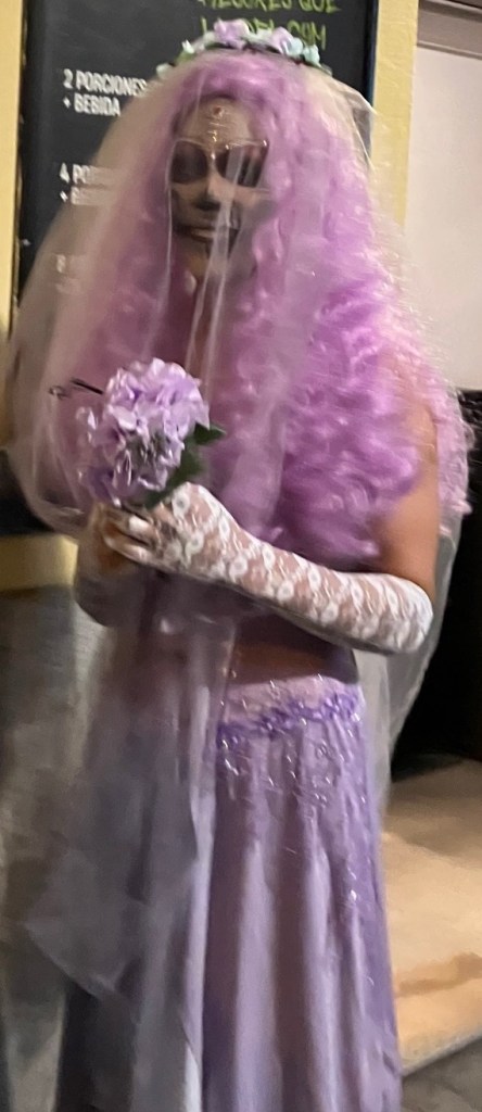 photo of a person in skull-face makeup and sunglasses, wearing a long purple wig, purple veil, glittery purple dress, lilac lace gloves, holding a purple bouquet
