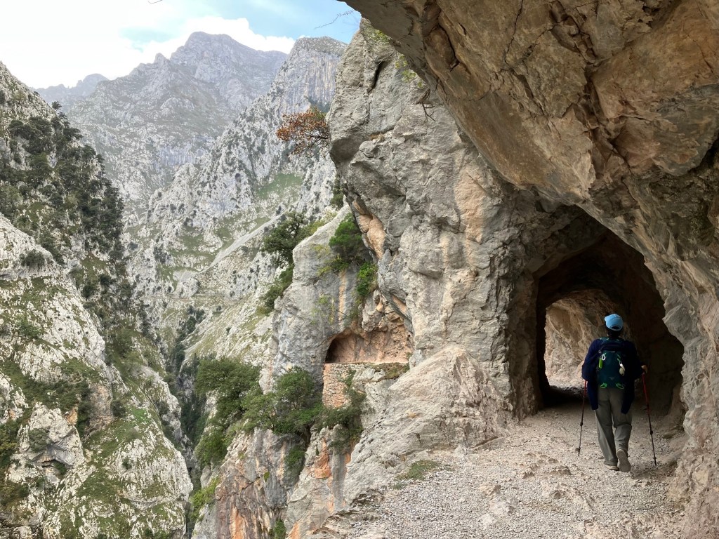 Photo of a person in a blue cap walking with hiking poles into a small tunnel carved out of a steep cliff, with jagged gray rock peaks and cliffs plunging off the side.