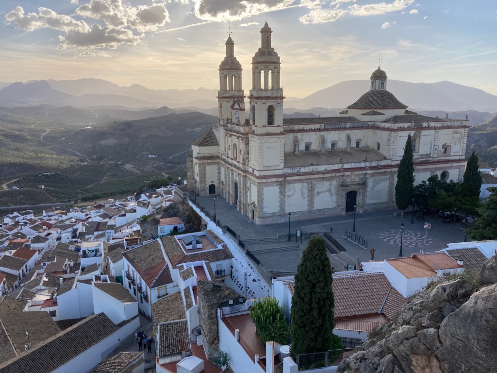 Photo of a beige stone cathedral with two spires and a dome atop a hill with white-walled houses spread across the hillside beneath it. In the background are mountains and the sun gleaming through clouds.