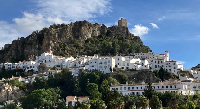 Photo of a cliff-sided hill with a rectangular, stone castle tower on top. Below the cliff is a village of white walled houses set on a hill amid green trees. Blue sky and white clouds are above.