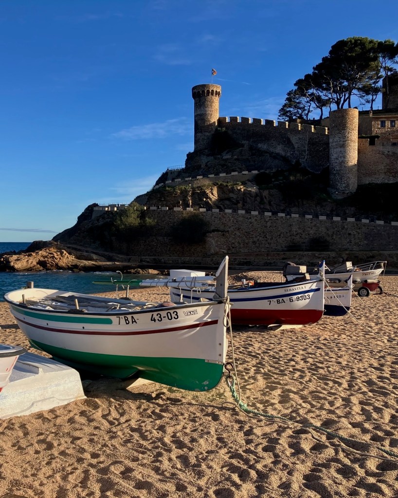 photo of 5 small, open-cockpit, wooden fishing boats lined up on a sandy beach, with a castle wall and towers in the background against a blue sky