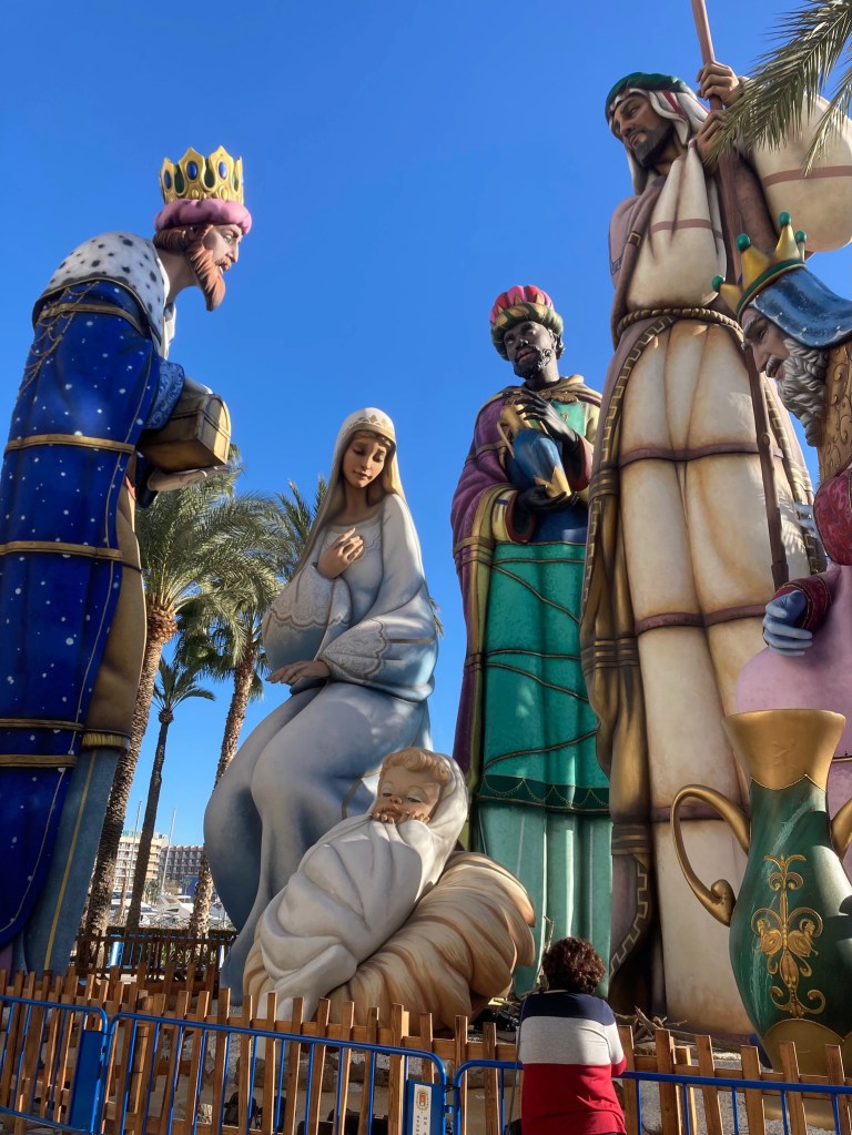 photo of the three magi, Mary, and baby Jesus, all towering above palm trees in the background and a person in the foreground who seems miniature by comparison