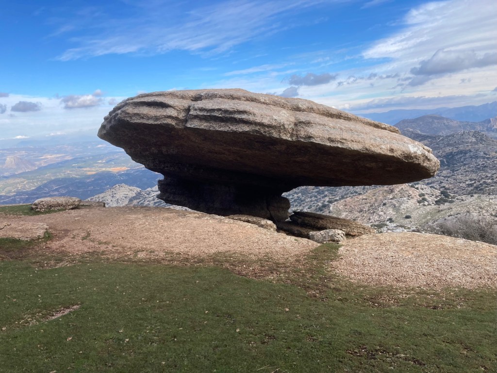 photo of a wide flat grey rock shaped like a pommel on a saddle, with a small neck supporting it on a flat rocky ledge overlooking a wide valley.