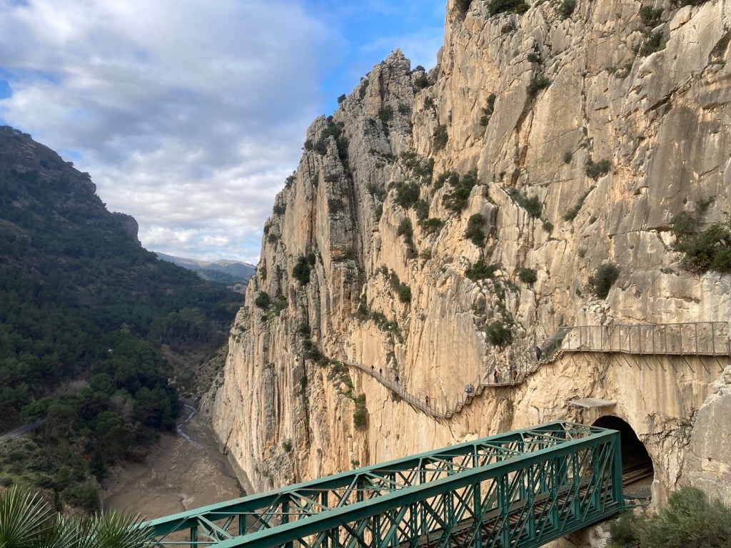 photo of a tan vertical cliff next to a dry river gorge with a green trestle bridge entering a tunnel at the bottom of the image, halfway up the cliff, and a tiny walkway with a small fence attached to the cliff.