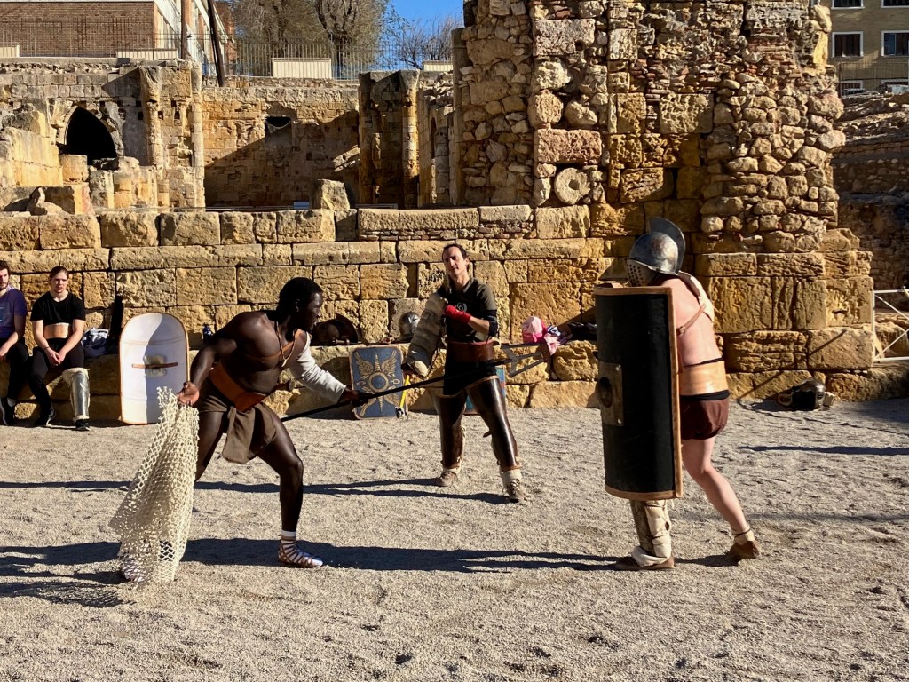 photo of a black man holding a net and a spear, crouched facing a white man in an iron helmet, holding a brown shield. A third man stands in between as judge. Behind them is a sand-colored wall of massive old bricks.