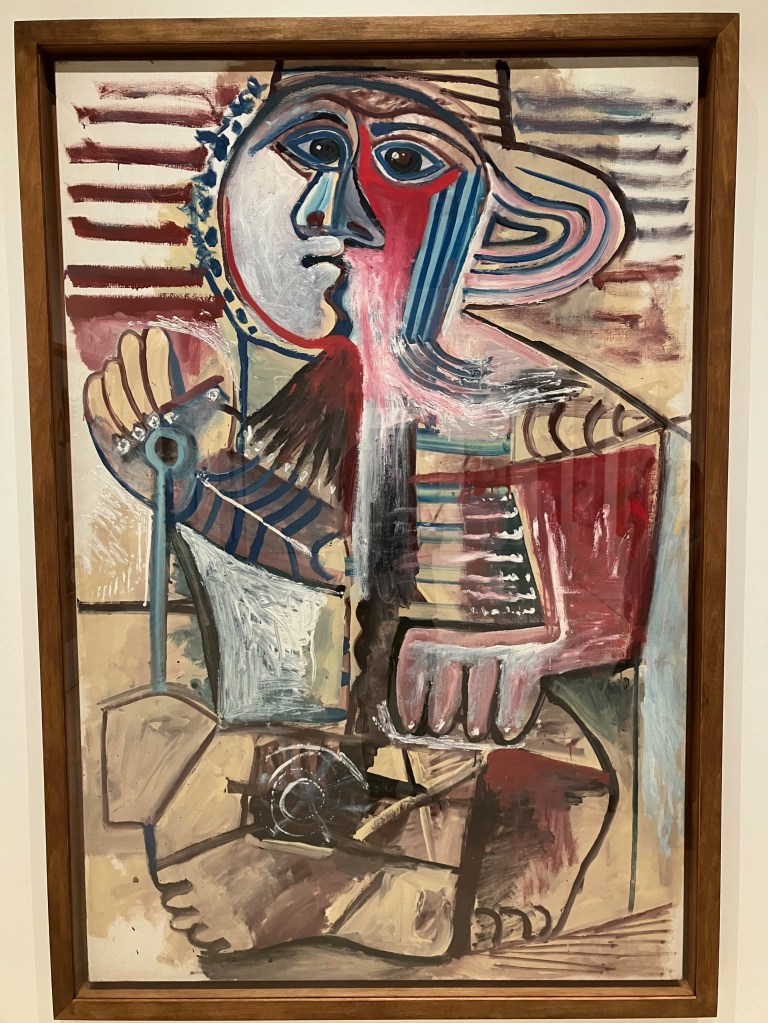 photo of an abstract painting of a child in brown, black, red, and blue, with a face at the top, a hand holding a sand shovel, and legs curled underneath the body.