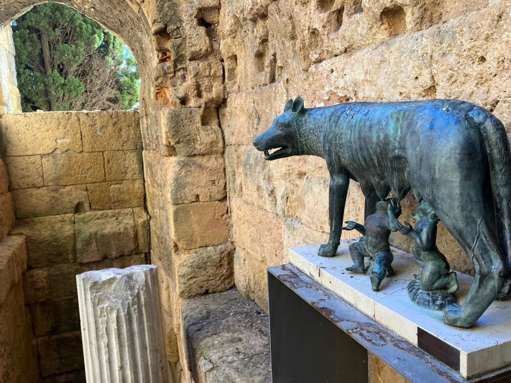 Photo of a bronze replica of the famous statue depicting the myth of Rome's founding -- Romulus and Remus with a she-wolf -- standing guard by the 2nd Century BC Roman wall and a fragment of a column.