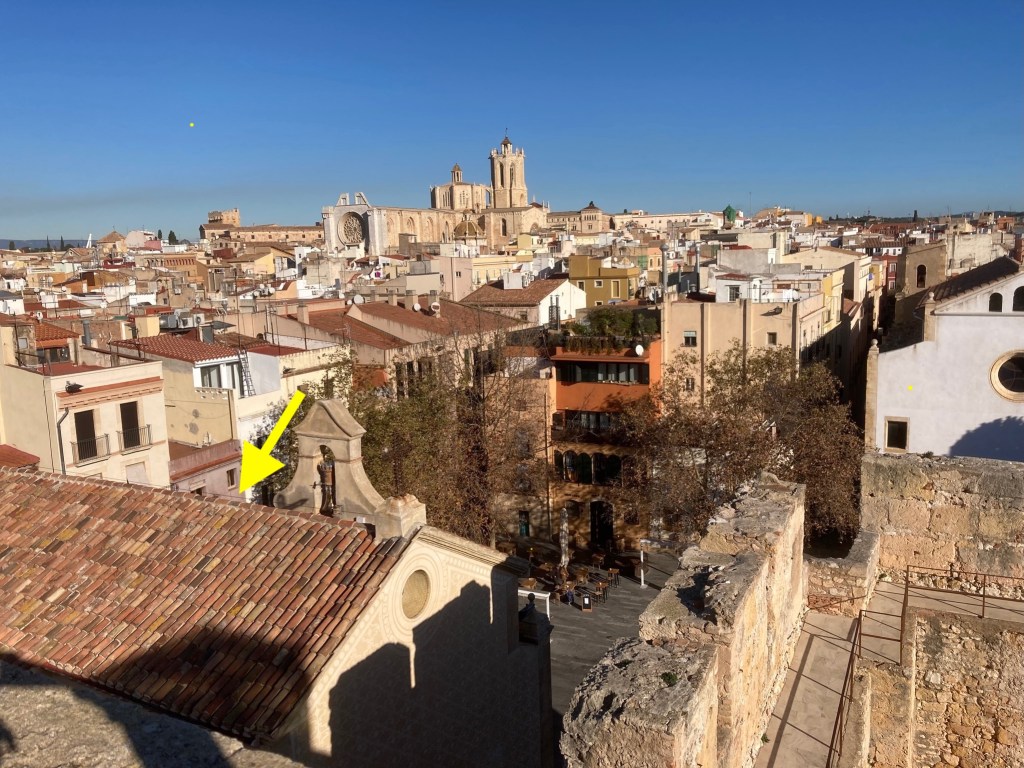 photo of a sandy brick wall pointing across a courtyard to a row of buildings, overlooking red and brown rooftops with a cathedral on the horizon. A yellow arrow points to one of the buildings around a courtyard far below.