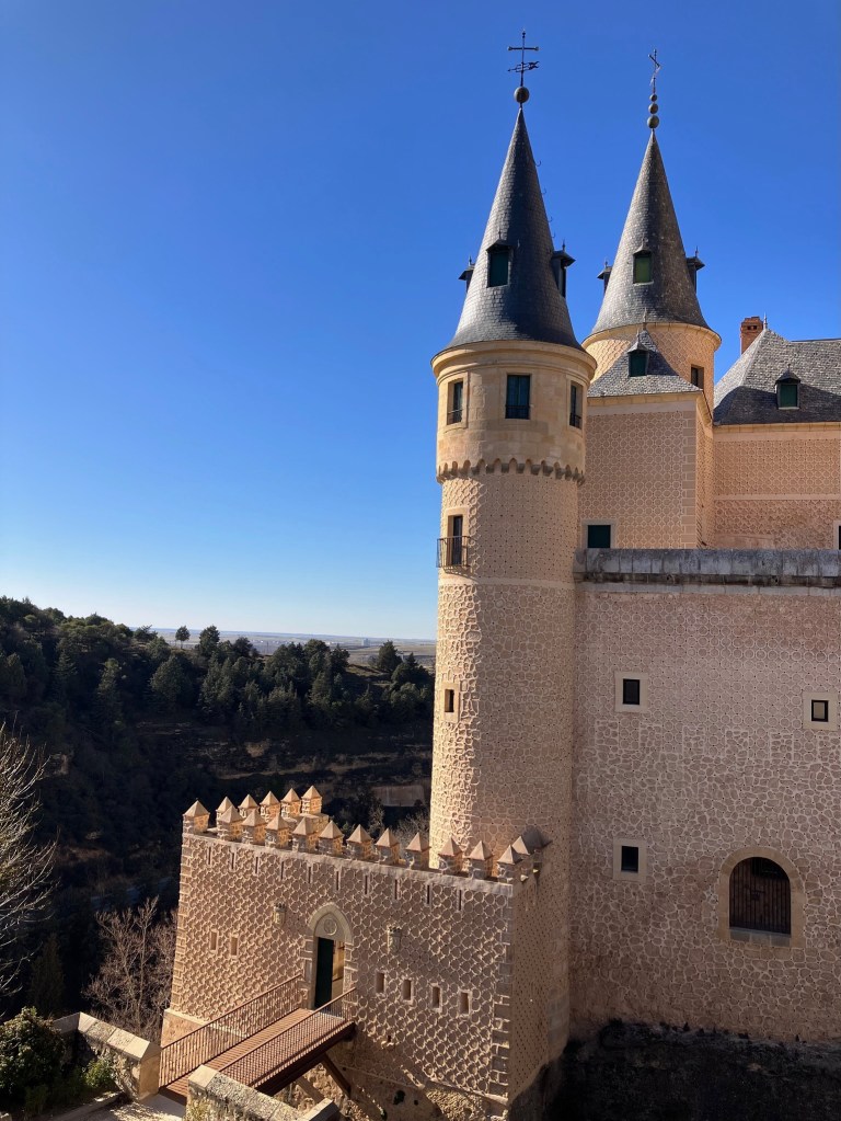 photo of a light brown castle with two tall, cylindrical towers with pointed gray slate roofs and a succession of walls framed against a bright blue sky