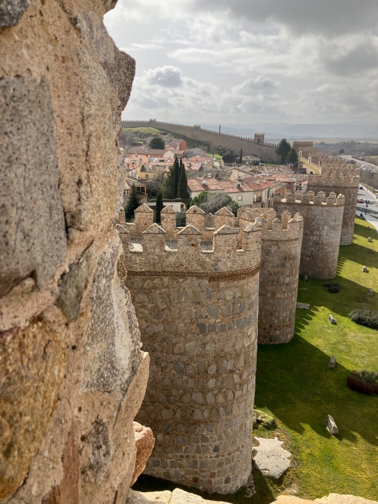 photo of semi-circular sandstone towers along a wall around a town with red-roofed houses inside and green grass outside