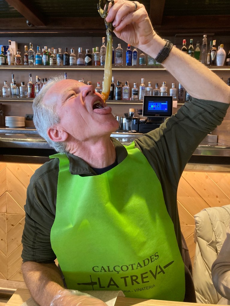 photo of a white man wearing a green bib that says "Calcotades" holding a cooked leek dipped in orange sauce above his open mouth with three rows liquor bottles behind him in a restaurant.