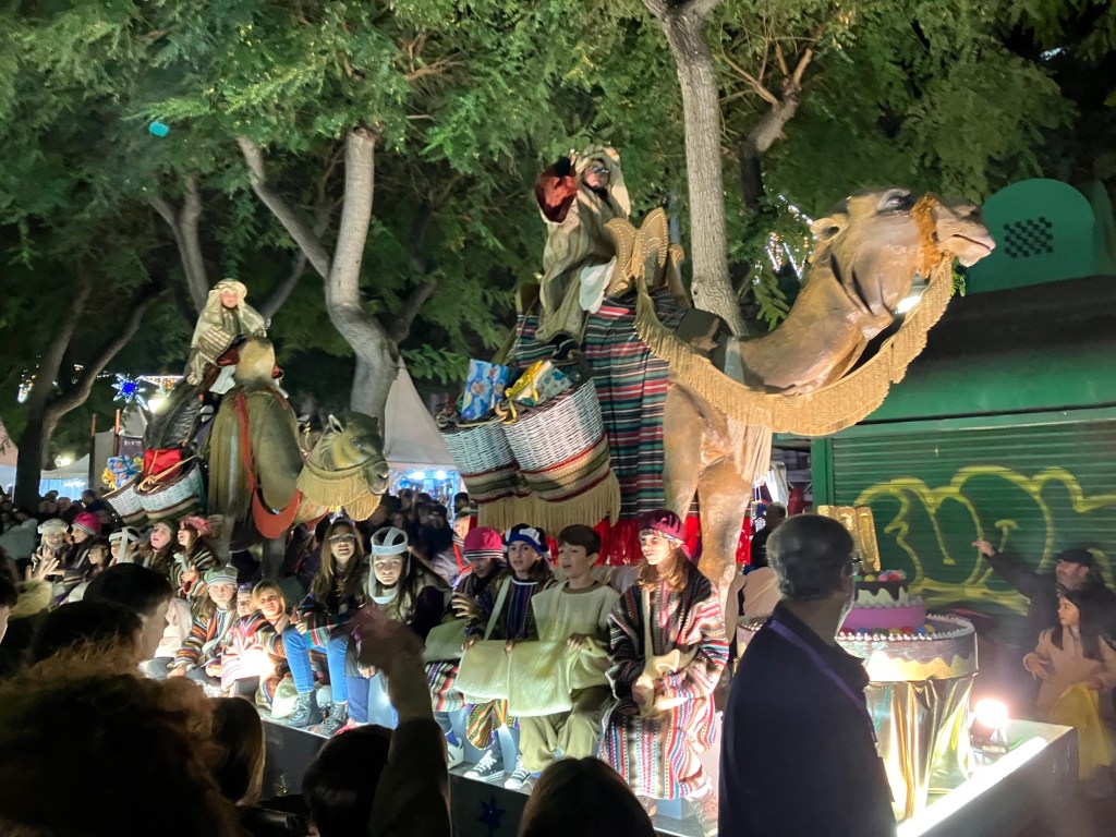 Photo of a parade float with two large camels draped in blankets and hanging baskets filled with gifts, ridden by two children dressed as desert travelers